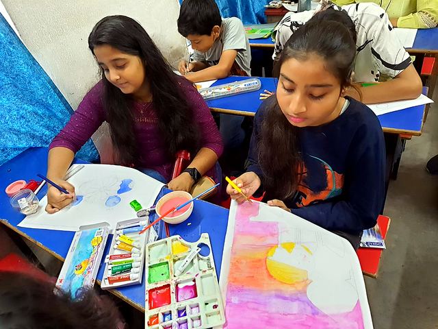 Children painting on sheets with water colour boxes on the table